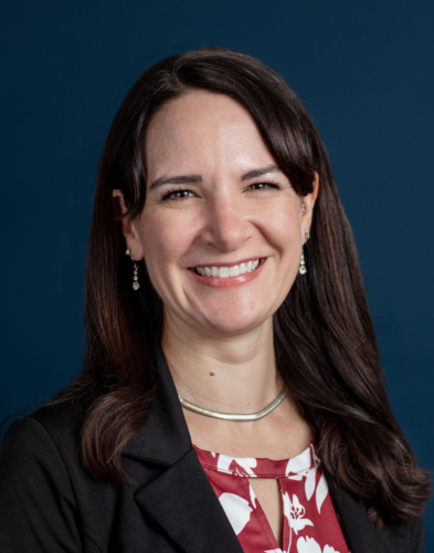Woman with long brown hair wearing a black blazer and red and white floral blouse, smiling in front of a dark blue background.